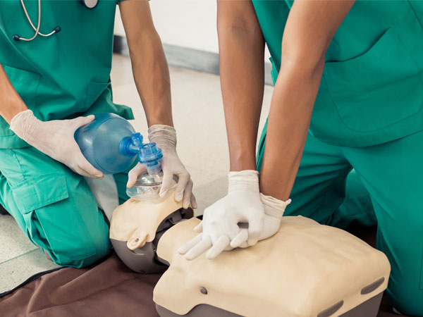 A picture of a CPR dummy. There is one person standing over it performing CPR and the other is regulating breathing using a ballon-ish device (name pending)