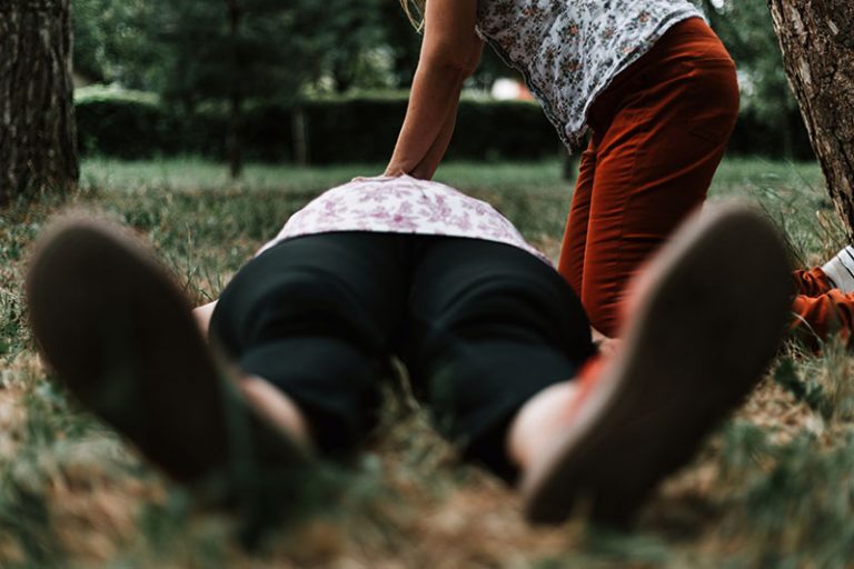 A person is lying on the ground with their feet facing the camera. There is another person standing over them with their hands on the first persons chest, they appear to be performing CPR