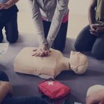 A group of people kneeling around a CPR dummy with a first aid kit nearby. One of them is performing CPR.