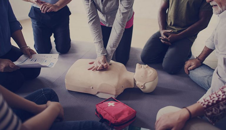 A group of people kneeling around a CPR dummy with a first aid kit nearby. One of them is performing CPR.