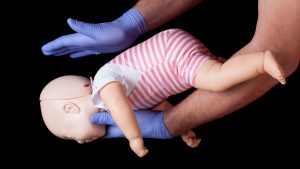 A baby held face down, by a man wearing surgical gloves. The man is holding on hand above the baby's back.
