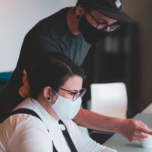 A picture of two people wearing masks, sitting at a table and pointing at something offscreen.