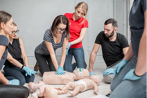 A picture of a group of people sitting besides an adult CPR doll and two baby CPR dolls . One of the people is performing CPR on the adult doll.