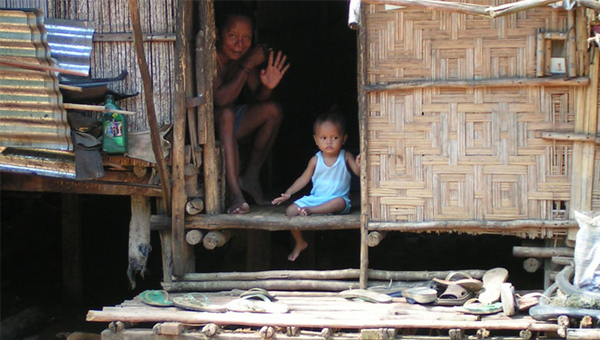 A woman and a child are sitting in the door of a woven house, the woman is waving at the camera and the child is distracted. There are multiple ordinary things strewn around the entrance