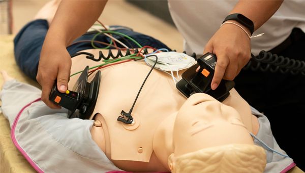 A person is kneeling over a CPR dummy, they are holding defibrillator on the dummies chest