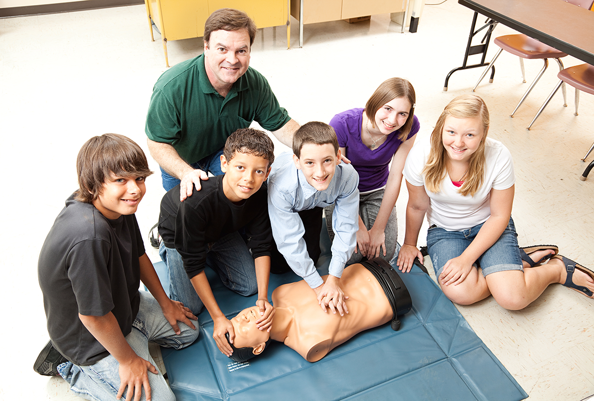 Six people are kneeling next to a CPR dummy, the center one has their hands on the dummies chest. There are all looking up at the camera